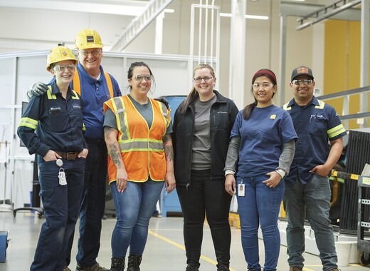 Six employés de SageGlass en vêtements de travail et lunettes de sécurité collaborant dans une usine de fabrication.