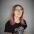 Headshot of a person wearing a black t-shirt and glasses and dyed pink long hair and glasses.