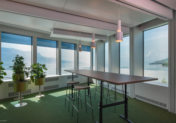 Intérieur de la salle de réunion avec table haute et vitres intelligentes donnant sur l'eau et les montagnes. 