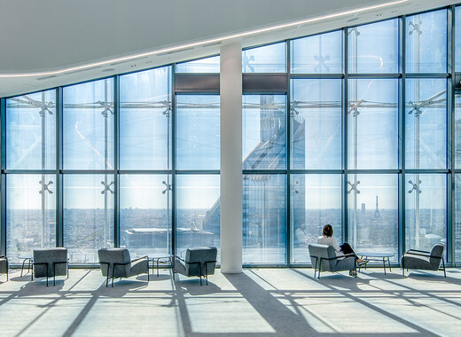 Espace commun intérieur d'un immeuble de bureaux avec, de gauche à droite, des chaises longues et des baies vitrées intelligentes. 