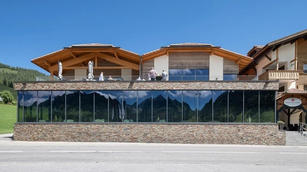 Vue extérieure d'un bâtiment avec du verre et de la pierre des champs en bas et une terrasse en haut lors d'une journée ensoleillée.